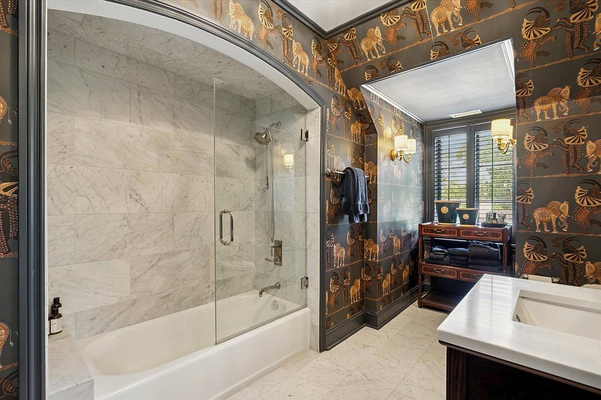 This bathroom features a striking combination of dark, patterned wallpaper depicting stylized animals and a light-toned stone shower surround. The space includes a built-in bathtub with a glass panel, a vanity with a stone countertop, and a shelving unit for storage. The perspective is from the doorway, looking into the room to showcase the contrast between the bold wall treatment and the neutral tile flooring.