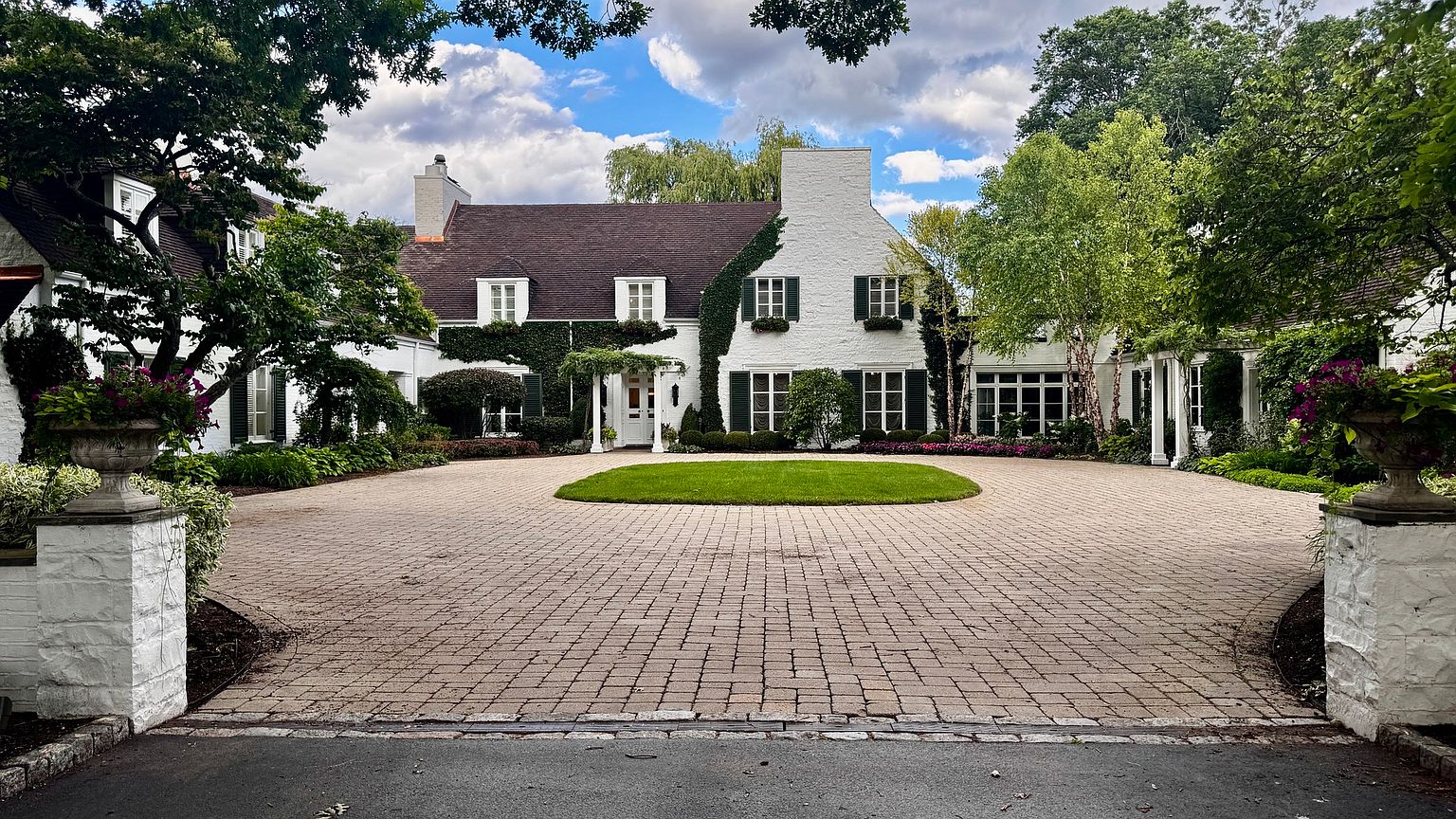 This stately white-painted brick residence features a classic architectural style with a symmetrical facade, dark shutters, and lush ivy climbing the exterior walls. A large, circular paver driveway centers around a manicured lawn, framed by mature trees and stone pillars at the entrance. The perspective is a wide, eye-level shot that captures the grand scale and inviting curb appeal of the property.