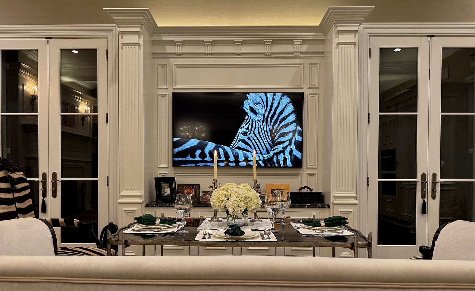 This elegant dining room features a formal table set for a meal, centered in front of a sophisticated built-in cabinetry unit with a television displaying a zebra image. The room is framed by symmetrical glass-paned doors on either side, creating a balanced and refined aesthetic. The space is illuminated by soft, warm lighting, including candles on the table and recessed lighting above the cabinetry, contributing to an intimate and luxurious atmosphere.