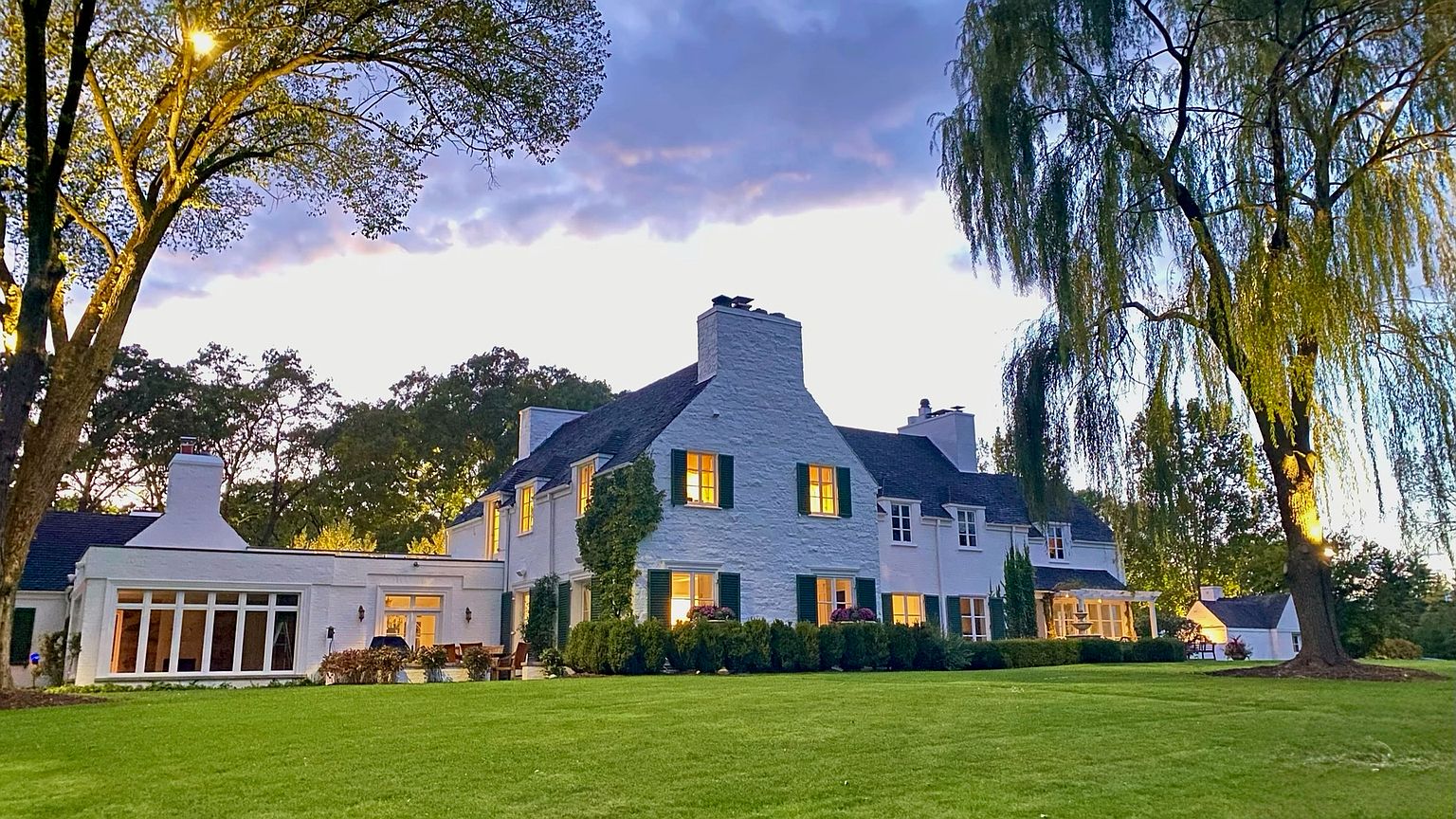 This expansive rear view captures a stately white-painted stone residence set against a twilight sky. The home features a multi-gabled roofline, symmetrical window placement with dark shutters, and a sprawling, well-manicured lawn that leads up to the structure. Large, mature trees frame the property, enhancing the serene and established atmosphere of this classic estate.