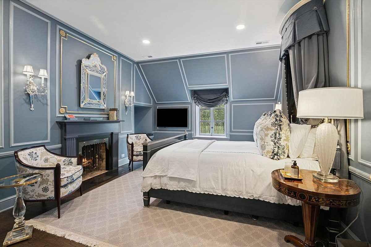 This elegant primary bedroom features sophisticated blue paneled walls with gold trim and a classic fireplace with a decorative mirror above. A plush bed with white linens and patterned pillows serves as the focal point, complemented by a round side table and a patterned armchair. The room is bathed in soft light from wall sconces and a bedside lamp, creating a refined and tranquil atmosphere.