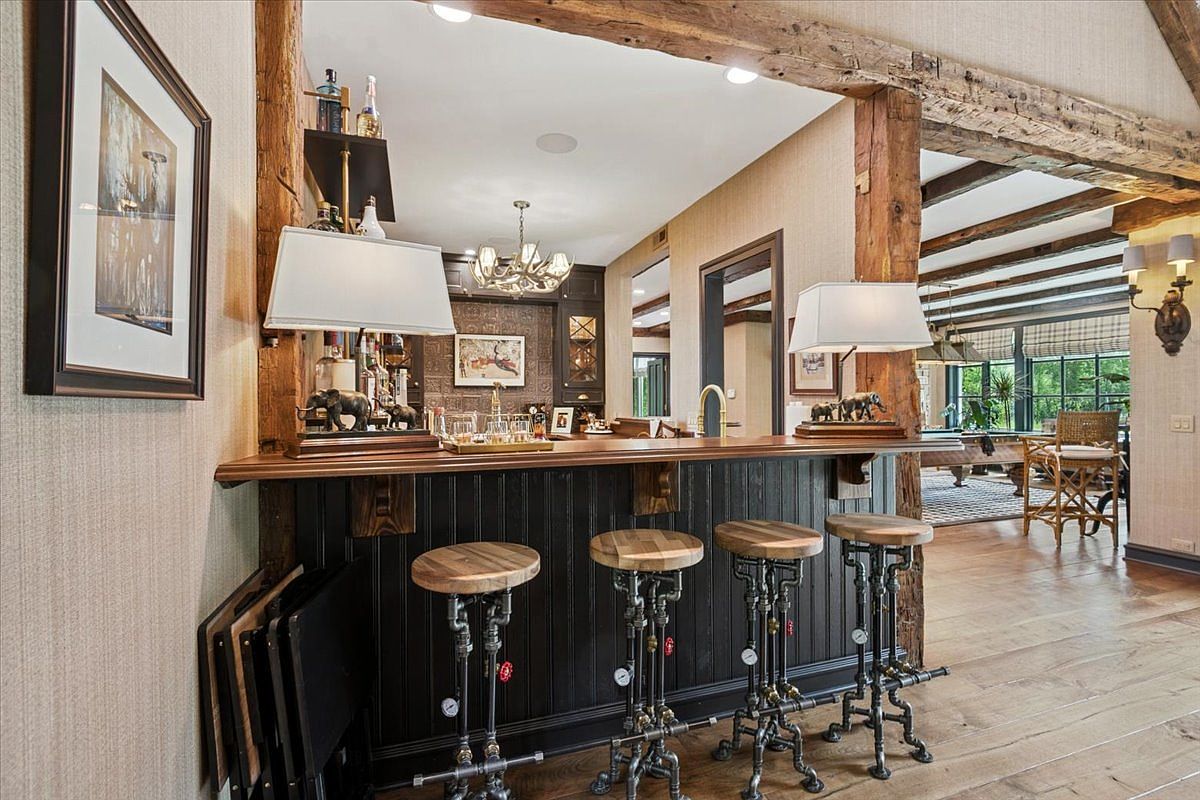 This image showcases a rustic-style home bar area featuring a dark wood counter with beadboard paneling and four industrial-inspired stools made of metal pipes and wood seats. The bar is framed by exposed timber beams, and the back bar area includes shelves with glassware and a decorative antler chandelier. The perspective looks directly at the bar, with a glimpse into an adjacent room containing a pool table and wooden furniture, creating a warm and inviting entertainment space.