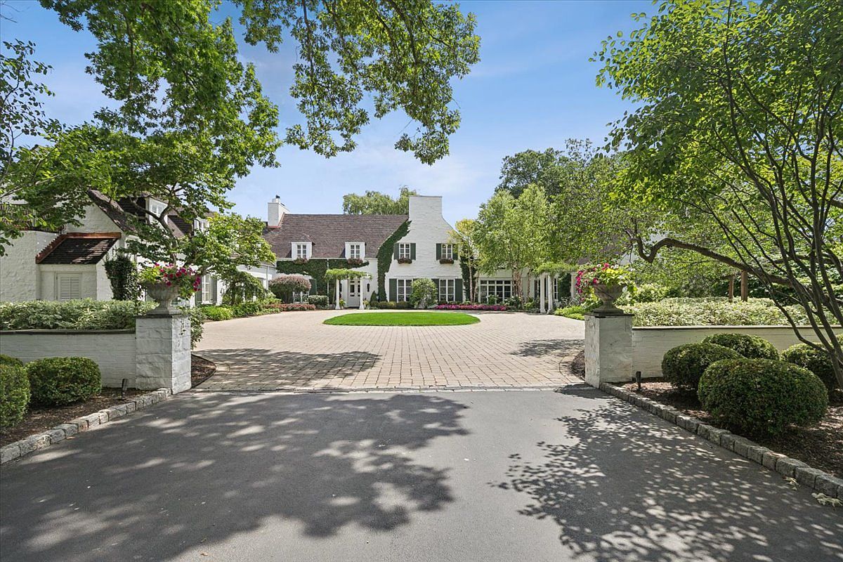 This elegant estate features a classic white-painted brick facade with climbing ivy and a symmetrical design. A circular paved driveway leads to the main entrance, framed by lush green trees and manicured landscaping. The perspective is a wide, eye-level shot capturing the grand arrival experience of the property.