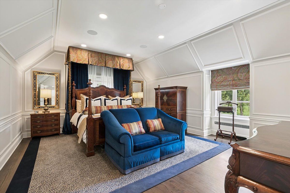 This elegant primary bedroom features a classic four-poster bed draped with rich blue curtains and a matching upholstered loveseat positioned at the foot of the bed. The room is characterized by sophisticated wall paneling, a patterned area rug, and a warm, traditional aesthetic enhanced by a wooden armoire and a matching dresser with a gold-framed mirror. Large windows with patterned Roman shades allow natural light to fill the space, creating a refined and comfortable atmosphere.