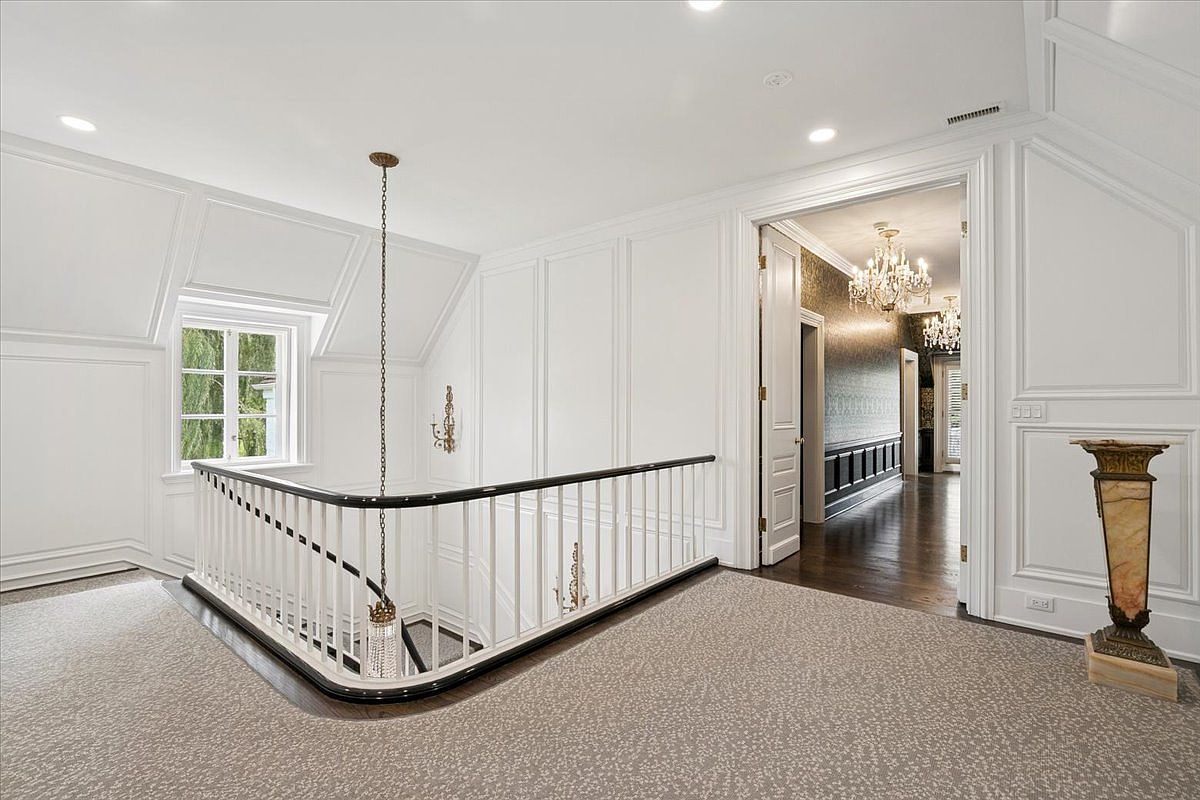 This elegant landing area features a curved staircase with a dark wood handrail and white balusters, overlooking a lower level. The space is finished with crisp white wall paneling and a neutral patterned carpet, leading into a hallway with dark wood flooring and a crystal chandelier. The perspective is from the landing, capturing the transition between the bright, open stairwell and the more intimate, classic interior beyond.