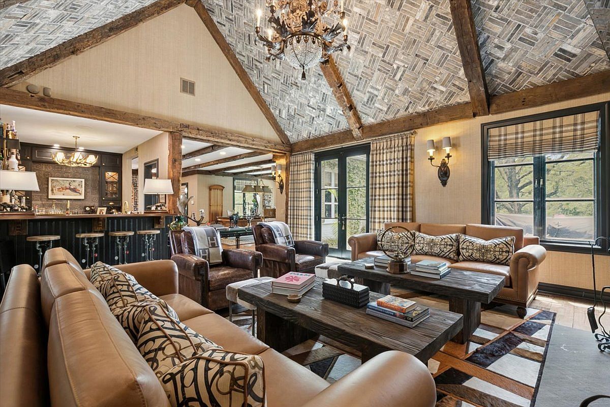 This expansive living room features a dramatic vaulted ceiling with exposed timber beams and a unique herringbone-patterned wood finish. The space is furnished with plush leather sofas and rustic wooden coffee tables, anchored by a geometric patterned area rug. A bar area is visible in the background, and the room is illuminated by a grand chandelier and warm wall sconces, creating a sophisticated, lodge-inspired atmosphere.