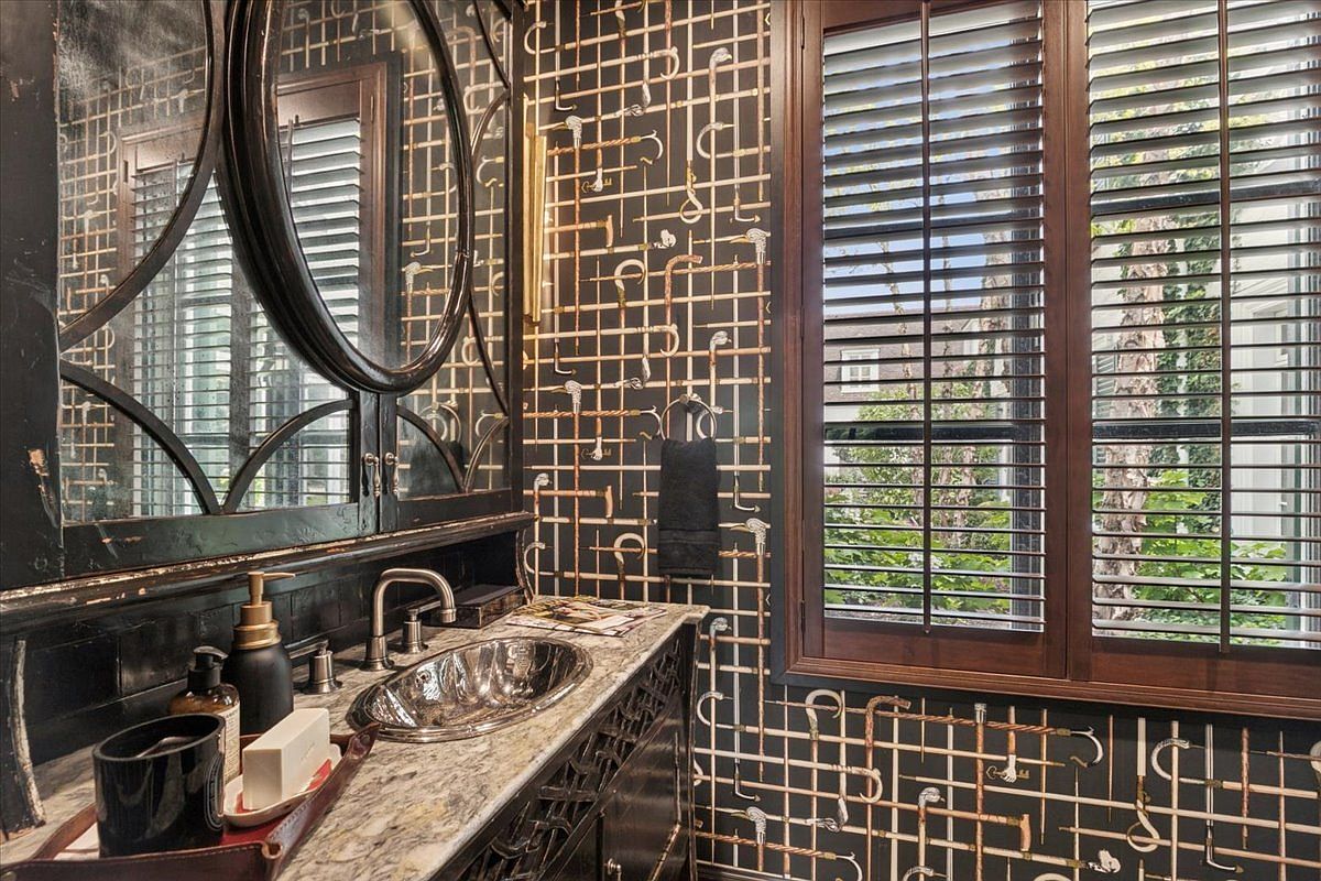 This powder room features a striking, eclectic design with patterned wallpaper depicting a grid of pipes and hooks. A dark, ornate vanity with a stone countertop and a metallic vessel sink serves as the focal point, complemented by a large, decorative mirror. The space is finished with dark wood shutters on the window, creating a moody and sophisticated atmosphere.