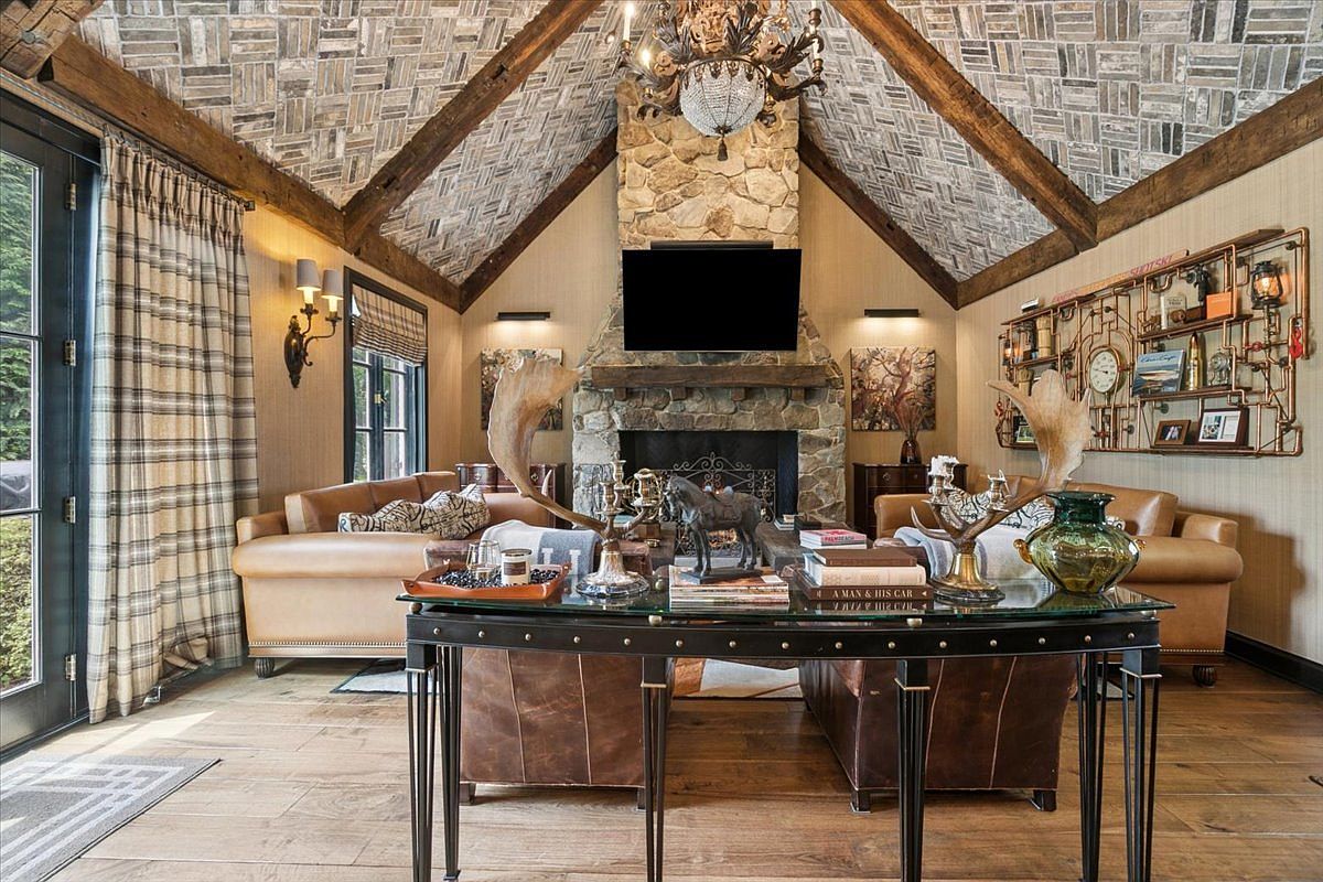This rustic living room features a vaulted ceiling with exposed timber beams and a unique herringbone-patterned wood ceiling treatment. A prominent stone-clad chimney breast serves as the focal point, housing a television above a substantial wooden mantel. The space is furnished with leather sofas and a decorative metal console table, creating a warm, lodge-inspired atmosphere.
