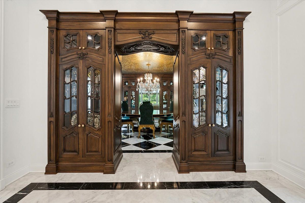 This image captures a grand transition space featuring a massive, ornate dark wood built-in unit that frames the entrance to a formal dining room. The cabinetry includes glass-paneled doors with intricate detailing, creating a sophisticated and traditional aesthetic. Beyond the archway, a glimpse of a dining area with a crystal chandelier and a patterned ceiling is visible, set against a backdrop of polished stone flooring with a dark geometric border.