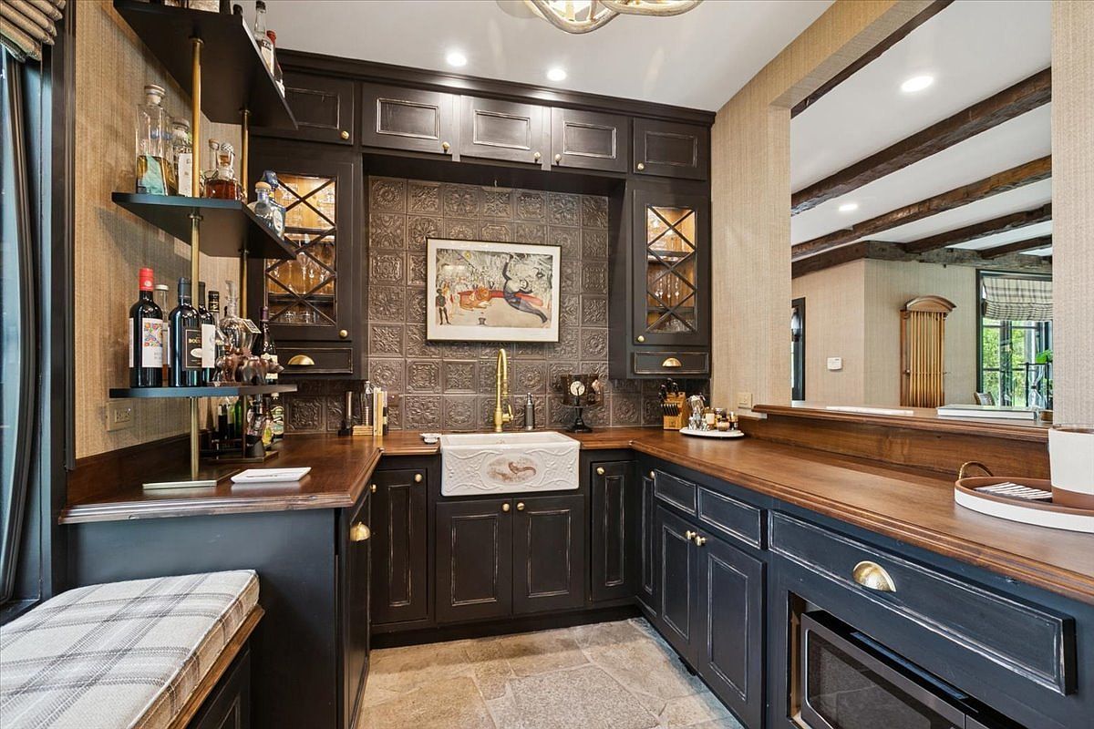 This sophisticated wet bar area features dark, custom cabinetry paired with rich, dark wood countertops and a distinctive patterned metal backsplash. A central farmhouse-style sink with a gold-toned faucet serves as the focal point, flanked by glass-front upper cabinets and open shelving stocked with glassware and bottles. The space is finished with stone flooring and a bench seat, creating an elegant and moody atmosphere perfect for entertaining.