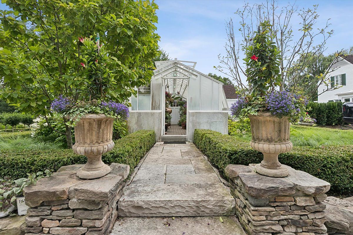 This charming garden scene features a stone-paved walkway leading directly to a classic glass-paned greenhouse. The path is framed by manicured boxwood hedges and two large, ornate stone urns overflowing with vibrant purple and green plantings. The perspective is centered and eye-level, highlighting the symmetrical landscaping and the serene, well-maintained atmosphere of the outdoor space.