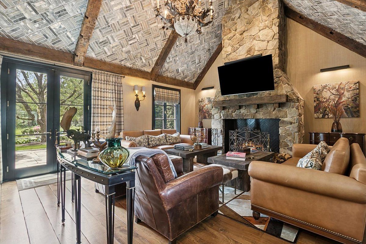 This rustic living room features a vaulted ceiling with exposed timber beams and a unique patterned tile finish. A prominent stone-clad chimney breast serves as the focal point, housing a fireplace and a mounted television above a wooden mantel. The space is furnished with comfortable leather seating, dark wood coffee tables, and a glass-topped console table, all set against a backdrop of warm-toned walls and large glass doors leading to the outdoors.