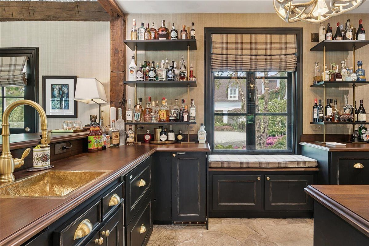 This sophisticated home bar area features dark, distressed cabinetry paired with rich wood countertops and a hammered metal sink. Open shelving displays an extensive collection of spirits, while a window seat with plaid cushions offers a cozy view of the outdoors. The space is accented by warm lighting and rustic architectural elements, creating an inviting and upscale atmosphere.