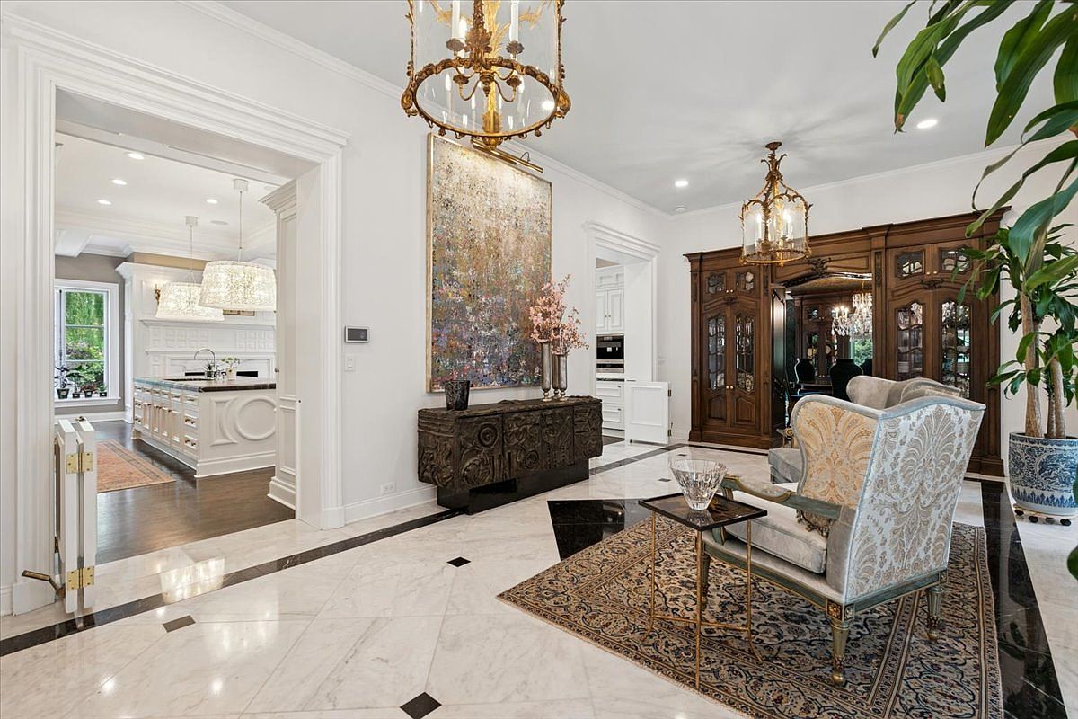 This elegant foyer features polished stone flooring with dark inlay accents and a grand chandelier centered above a patterned area rug. A large, textured abstract painting hangs above a dark, ornate console table, while a pair of upholstered wingback chairs creates a sophisticated seating area. The perspective looks through a wide, arched doorway into a bright kitchen space, showcasing the home's open and luxurious floor plan.
