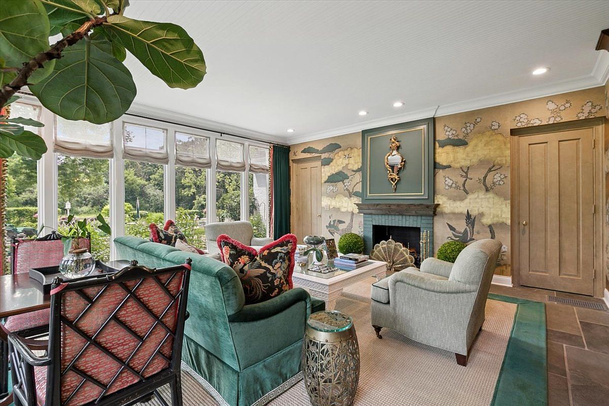 This bright sunroom features a wall of large windows that flood the space with natural light and offer views of the surrounding greenery. The room is anchored by a fireplace with a dark green surround and a decorative mirror, set against a wall adorned with ornate, nature-inspired wallpaper. Furnished with a plush green velvet sofa and a light-toned armchair, the space exudes a sophisticated, classic aesthetic.