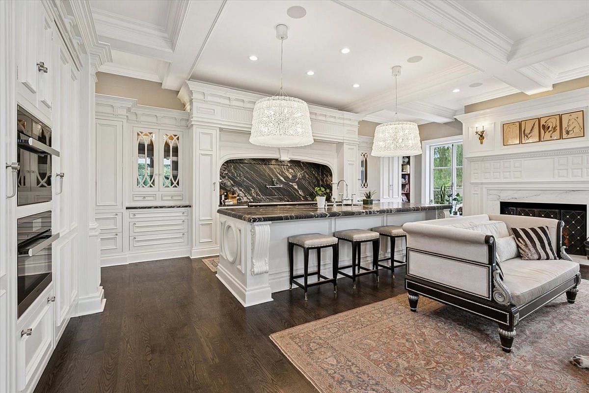 This expansive, high-end kitchen features white cabinetry, a large central island with a dark stone countertop, and three bar stools. The space is illuminated by two large, intricate pendant lights and recessed ceiling lighting, while a fireplace with a white mantel and a series of framed art pieces adds a cozy touch to the adjacent seating area. The room is anchored by dark wood flooring and a large, patterned area rug, creating a sophisticated and open-concept living space.