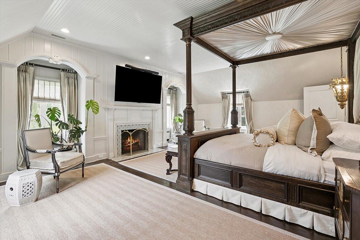 This elegant primary bedroom features a grand four-poster canopy bed as the central focal point, set against a backdrop of white paneled walls and a vaulted ceiling. A cozy seating area with a patterned armchair and a white ceramic stool sits near a fireplace with a classic mantel and a mounted television. The room is bathed in natural light from arched windows dressed in neutral curtains, creating a sophisticated and serene atmosphere.