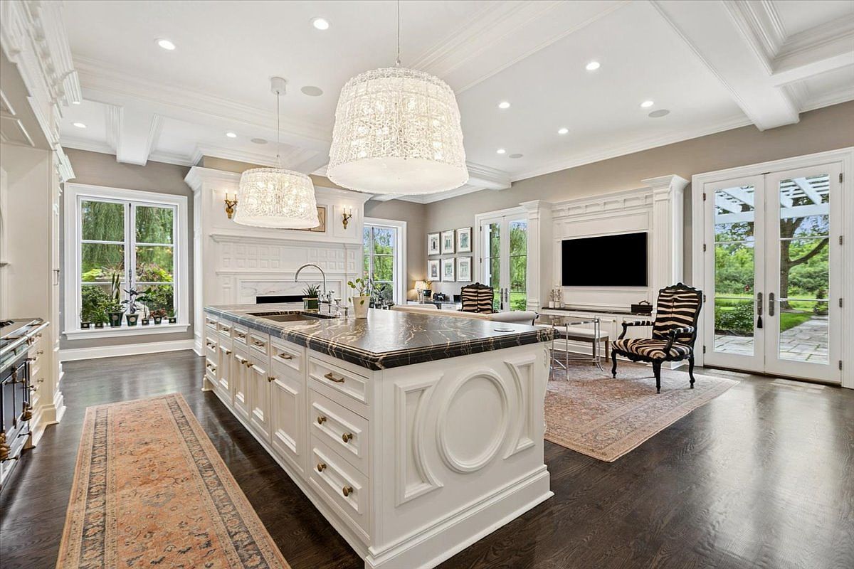 This expansive, open-concept kitchen features a large central island with a dark stone countertop and white cabinetry, illuminated by two prominent, textured pendant lights. The space flows seamlessly into a living area, showcasing dark hardwood floors, a wall-mounted television, and a zebra-print accent chair. Large windows and glass doors provide abundant natural light and views of the surrounding greenery, creating a bright and sophisticated atmosphere.
