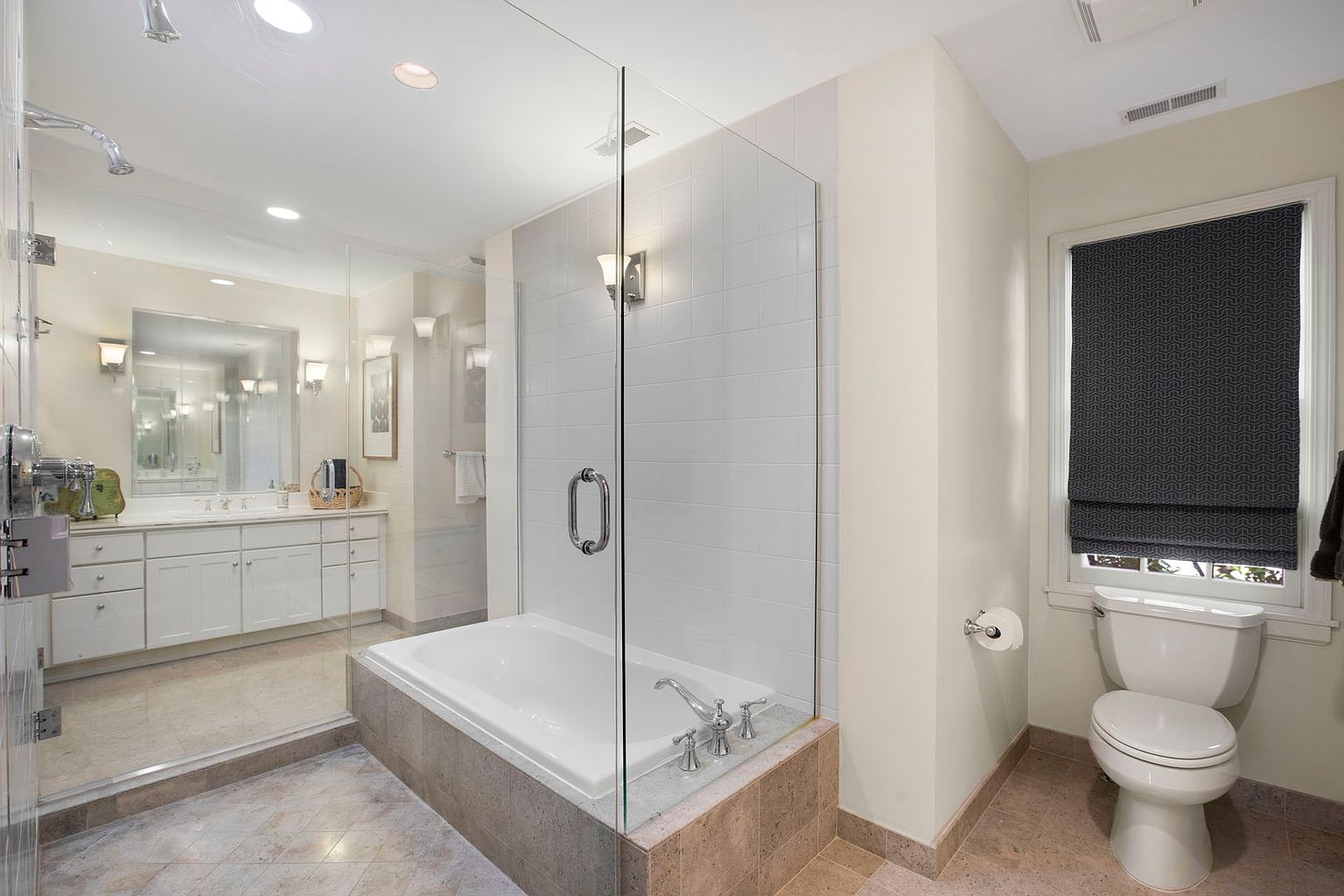This primary bathroom features a soaking tub enclosed by a glass partition, complemented by white tile walls and neutral-toned flooring. A white vanity with ample storage is visible in the background, while a toilet with a patterned window shade is situated on the right. The overall impression is clean, bright, and well-maintained.