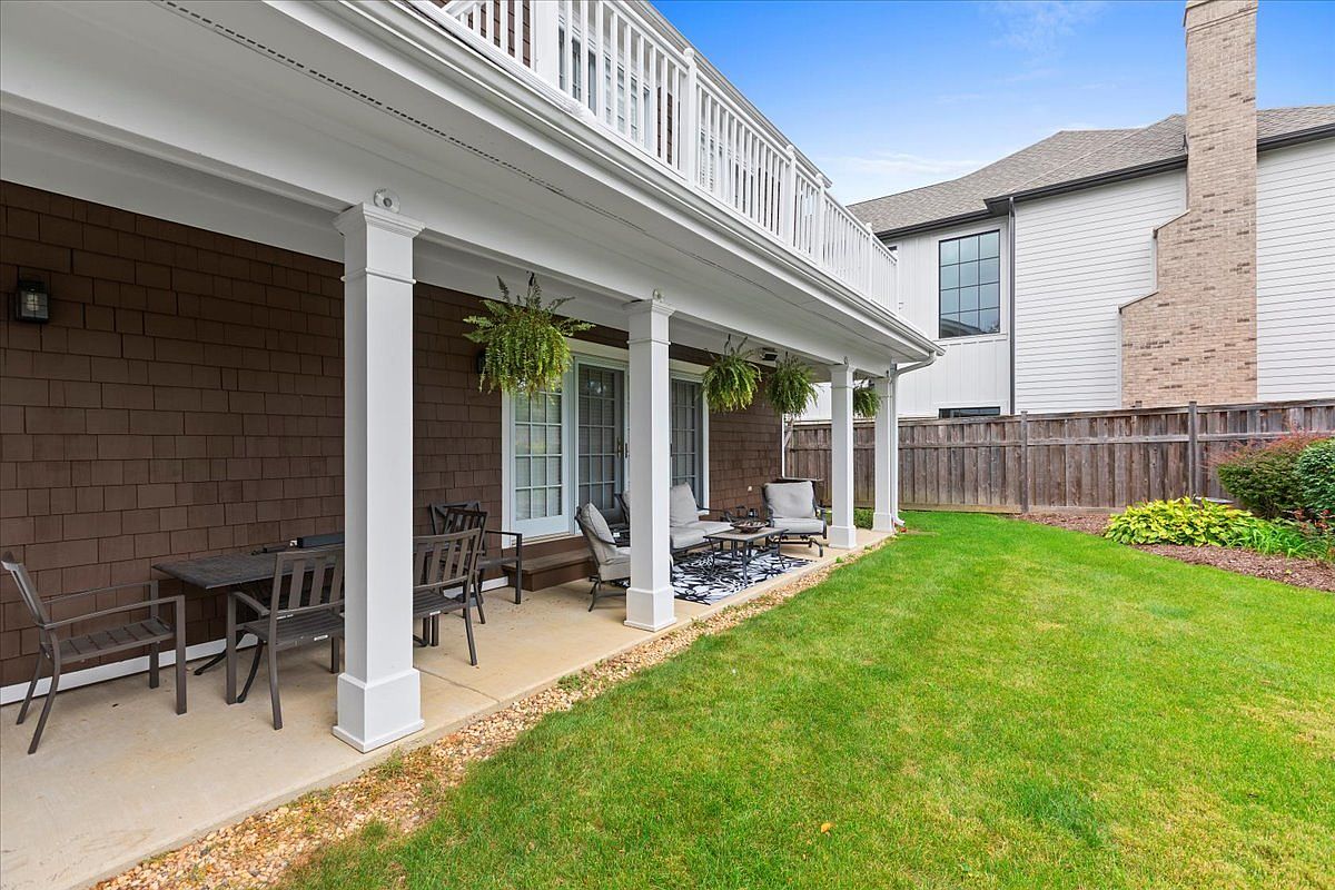 This image showcases a charming outdoor patio area, featuring a covered space with elegant white columns and a second-story balcony above. The patio is furnished with comfortable seating arrangements, including chairs and a small table, set upon a decorative rug. Lush green grass extends beyond the patio, creating a serene and inviting backyard setting.