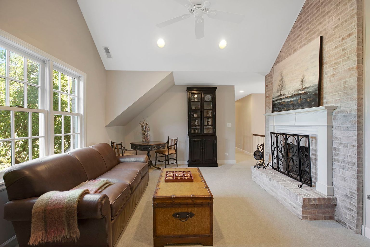 This is an interior shot of a living room featuring a brown leather sofa with a throw blanket, a trunk-style coffee table with a game on top, and a brick fireplace with a white mantel and decorative screen. A round table with chairs sits near a large window, and a dark wood cabinet is positioned against the wall. The room has a cozy and inviting atmosphere.