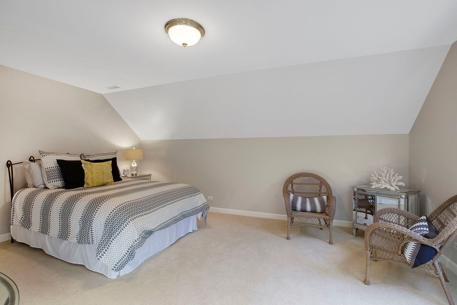 This is a cozy bedroom featuring a bed with a striped comforter and decorative pillows. The room has a neutral color palette with beige walls and light carpeting. Two wicker chairs and a mirrored side table with a decorative coral piece add to the room's charm and create a relaxing atmosphere.