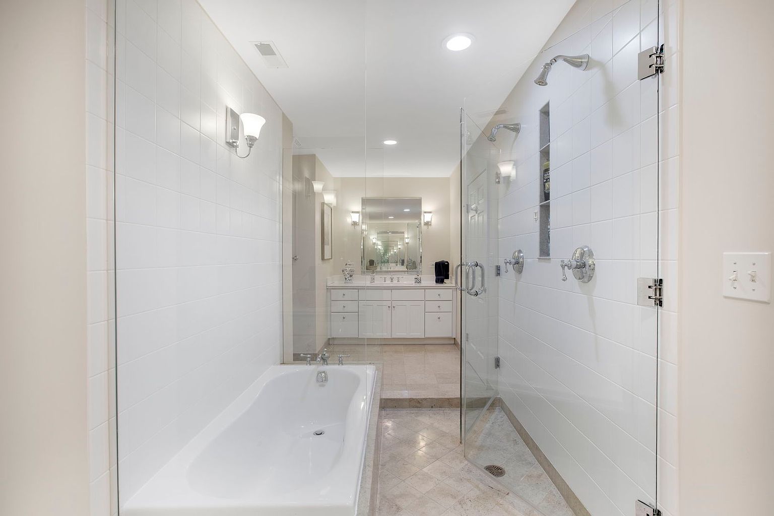 This is a bright and spacious primary bathroom featuring a soaking tub and a glass-enclosed shower. The bathroom has white tiled walls and a vanity with a large mirror and sconces. The flooring appears to be a light-colored tile, and the overall impression is clean and luxurious.