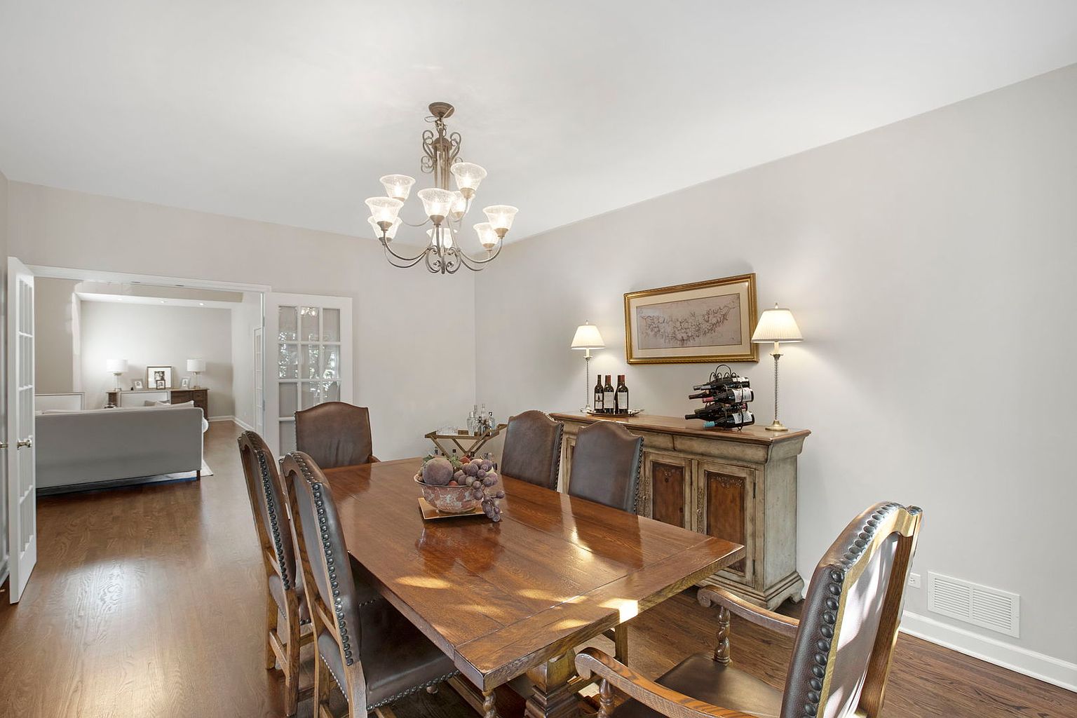 This is an interior shot of a dining room featuring a large wooden dining table with leather chairs. A decorative bowl of fruit sits on the table. In the background, there is a sideboard with lamps, wine bottles, and a framed picture, adding to the room's elegant and traditional style. A chandelier hangs above the table, providing warm lighting.
