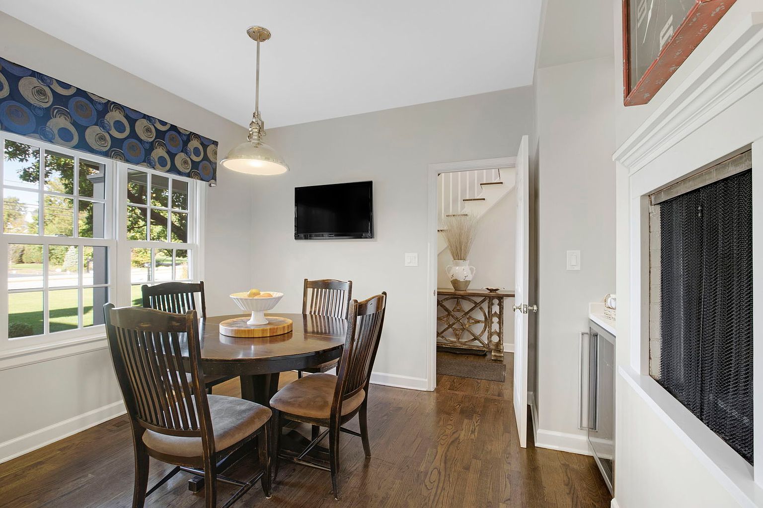 This is an interior shot of a dining room featuring a round wooden table with four chairs. A pendant light hangs above the table, and a window with a patterned valance provides natural light. A television is mounted on the wall, and an open doorway leads to a hallway with stairs, creating a sense of flow and connectivity within the home.