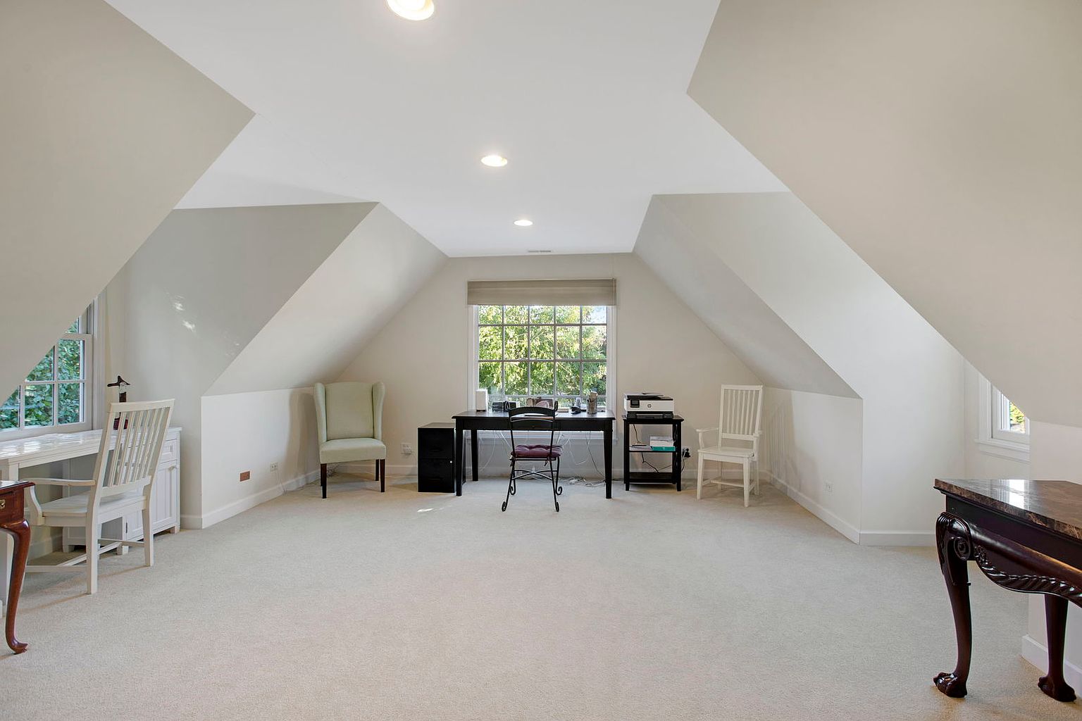 This is a spacious interior office/study featuring a neutral color palette and natural light. The room includes a desk setup with a chair, a filing cabinet, and additional seating options like a wingback chair and a white chair. The room has a carpeted floor and the walls are painted in a light neutral color, creating a bright and airy atmosphere.