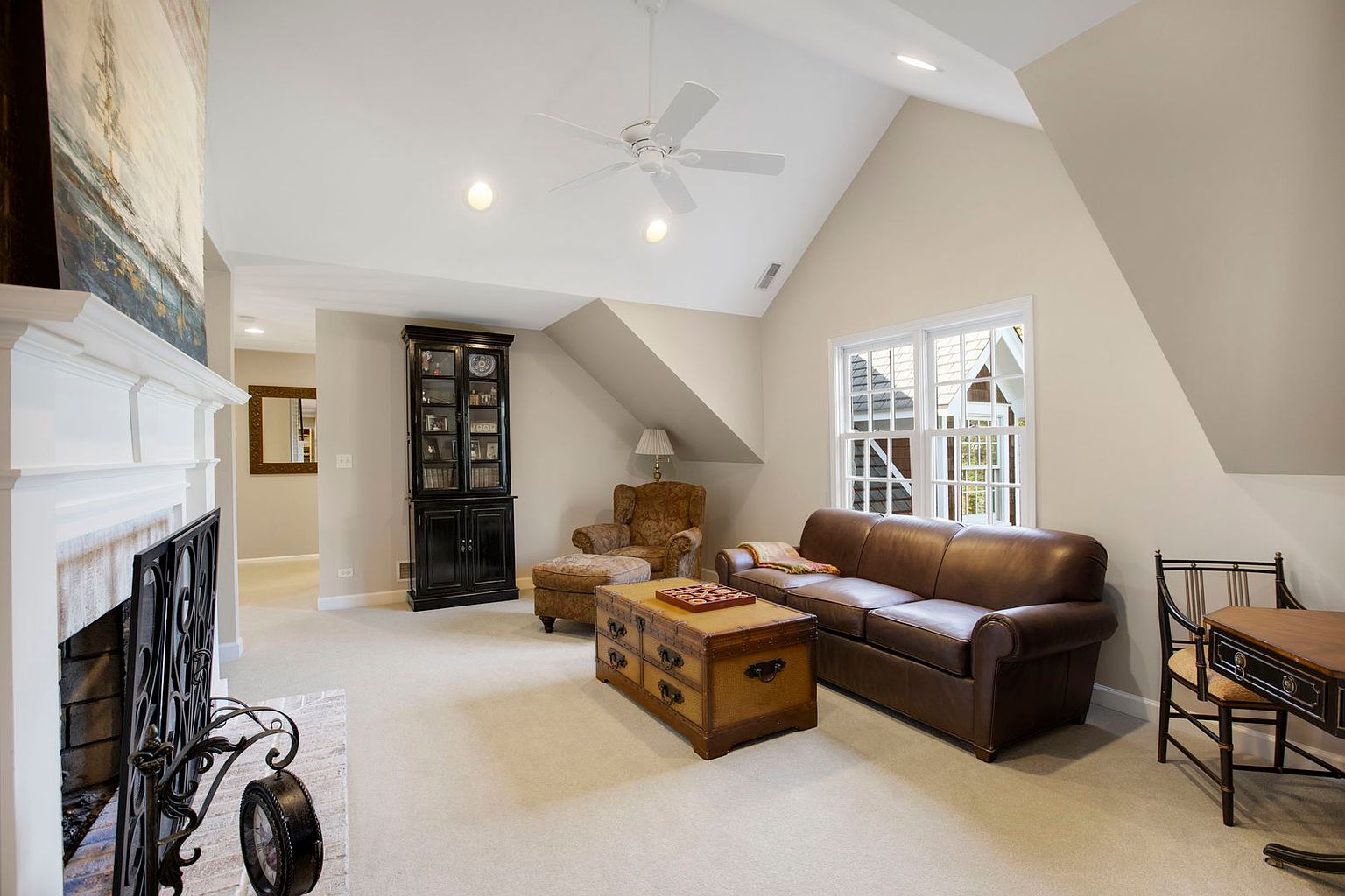 This is an interior shot of a living room featuring a brown leather sofa, a wooden chest-style coffee table, and a black bookcase. The room has a neutral color palette with beige walls and carpet, complemented by a white ceiling fan and trim. A fireplace with a decorative screen is visible on the left, and natural light streams in from a window on the right, creating a cozy and inviting atmosphere.