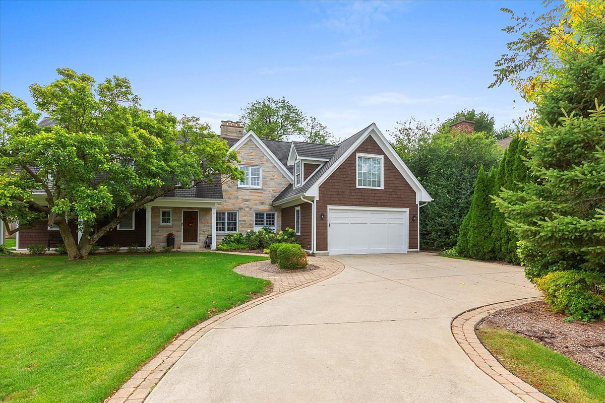 This is a front exterior view of a two-story house with a combination of stone and brown wood siding. The house features an attached two-car garage with a white door, a well-manicured lawn, and a curved driveway with brick edging. Mature trees and landscaping add to the property's curb appeal.