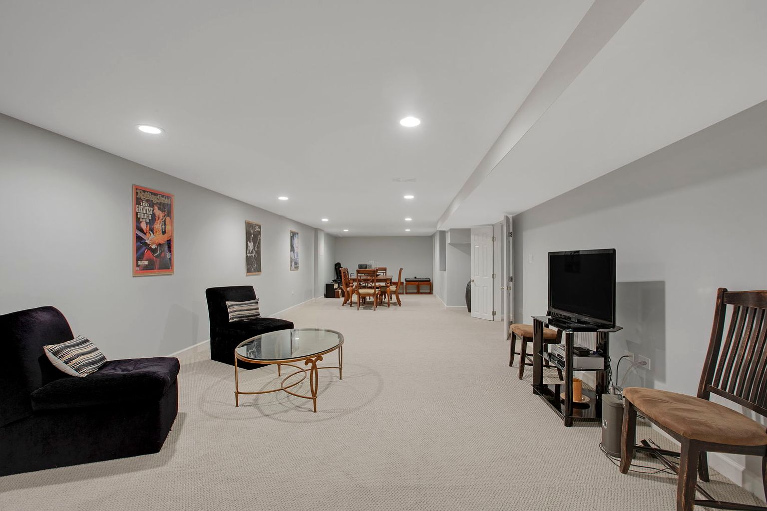 This is a spacious finished basement featuring neutral-toned carpeting and light gray walls. The area includes a seating area with dark armchairs and a glass-topped coffee table, a dining set in the background, and a television with a stand. The room is well-lit with recessed lighting, creating a comfortable and inviting atmosphere.