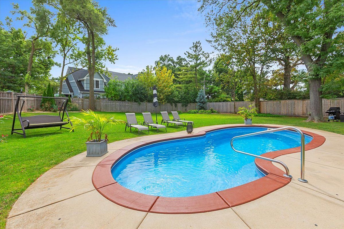 This image showcases a backyard pool area, complete with lounge chairs and a well-maintained lawn. The kidney-shaped pool features a blue interior and a red brick border, surrounded by a concrete patio. Lush greenery and trees provide privacy and a serene atmosphere, enhancing the property's appeal for relaxation and outdoor enjoyment.