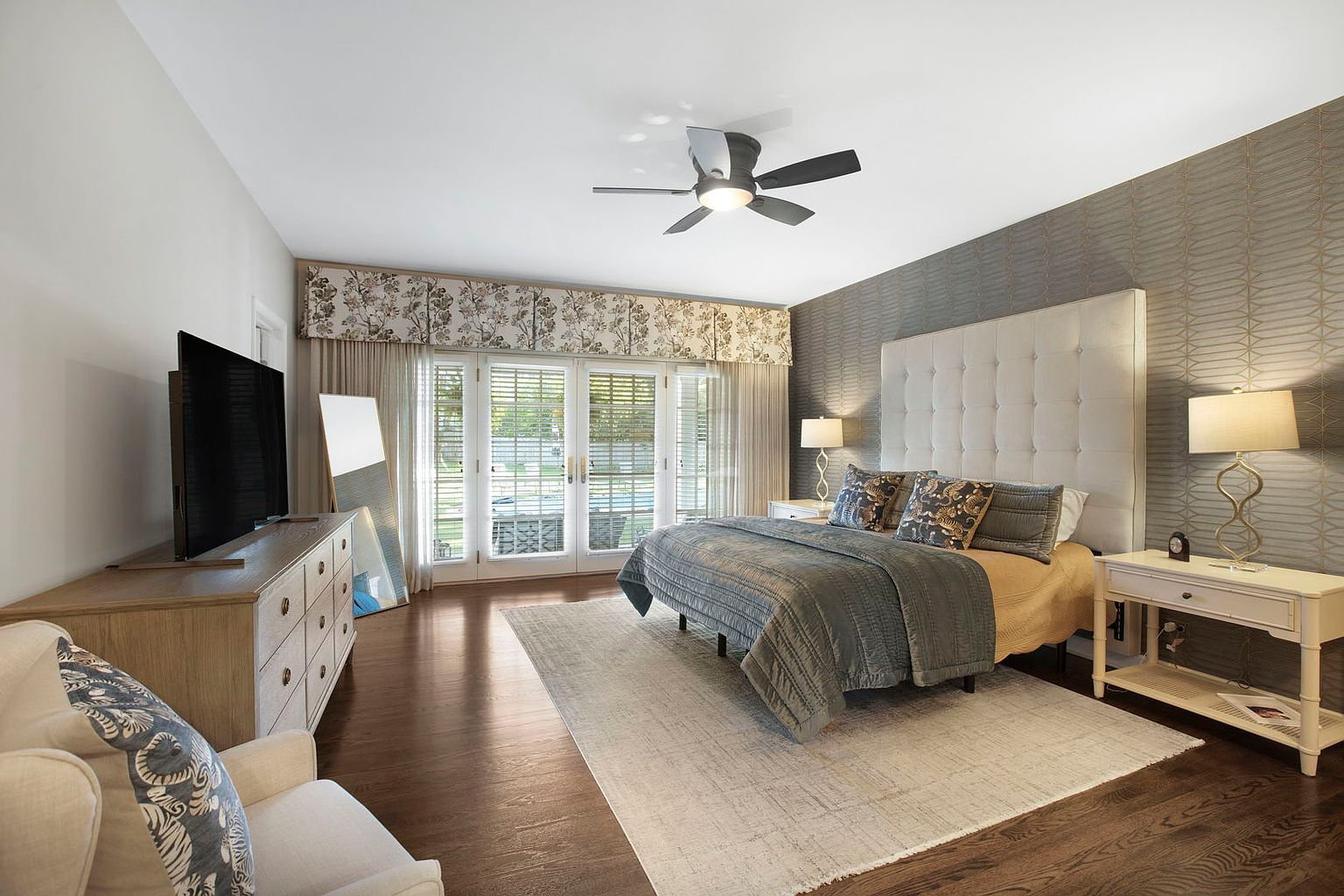 This is a primary bedroom featuring a large upholstered headboard, a bed with a gray quilt, and matching nightstands with lamps. The room has a neutral color palette with a textured wallpaper accent wall and dark hardwood floors. French doors lead to an outdoor area, providing natural light.