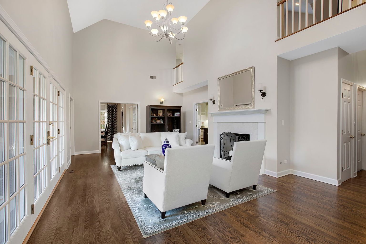 This is an interior shot of a living room featuring a high vaulted ceiling and hardwood floors. The room is furnished with a white sofa, two white armchairs, and a glass-topped coffee table, all arranged on a patterned area rug. A fireplace with a white mantel and a large mirror above it serves as a focal point, while a set of French doors provides natural light and access to another area.