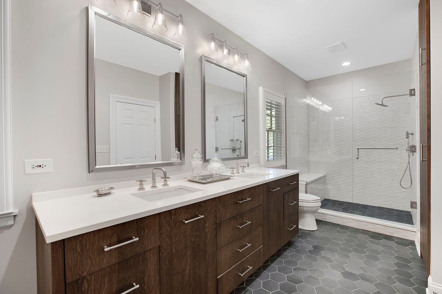 This is a well-lit primary bathroom featuring a double vanity with dark wood cabinets, white countertops, and modern silver fixtures. Two large mirrors hang above the sinks, reflecting the room's neutral color palette and the adjacent glass-enclosed shower with textured white tiles and dark gray flooring. The hexagonal floor tiles add a contemporary touch to the space.