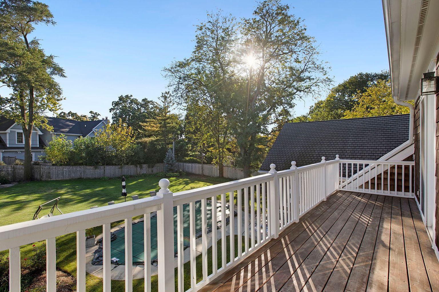 This image showcases a spacious wooden deck with white railings, offering a view of a well-maintained backyard. The yard features a covered pool, lush green lawn, and mature trees, creating a serene and private outdoor space. The sun shining through the trees adds a warm and inviting ambiance.
