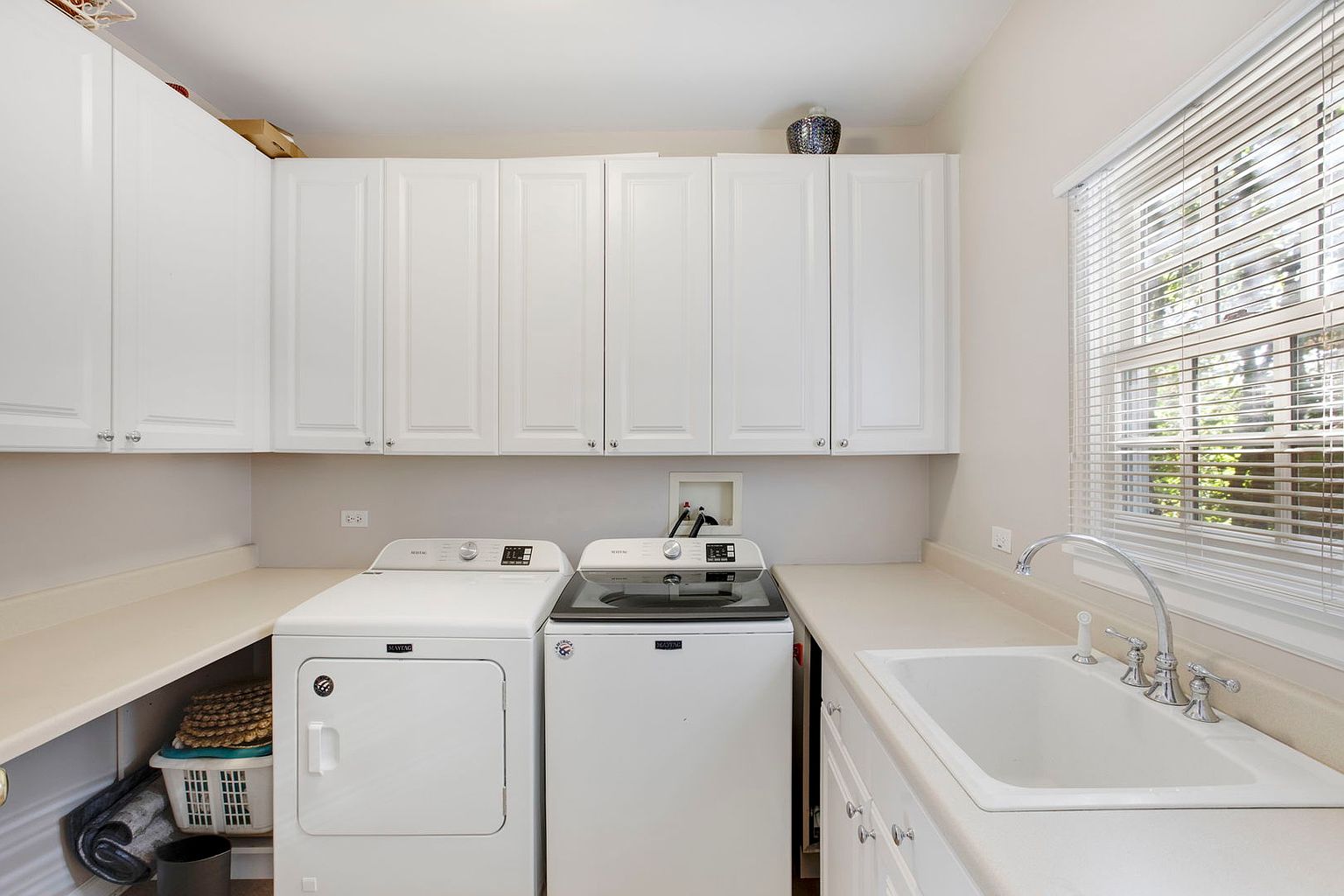 This is a well-organized laundry room featuring white cabinetry above a countertop that extends over the washer and dryer. A utility sink is integrated into the countertop, and a window with blinds provides natural light. The room appears clean and functional, ideal for a modern home.