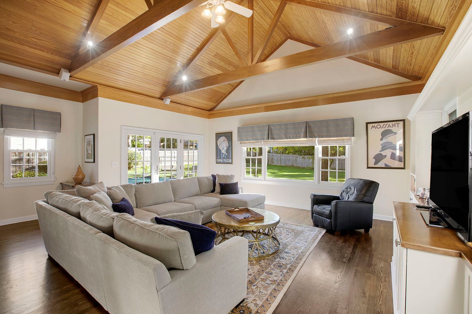 This is an interior shot of a living room featuring a large sectional sofa, a round coffee table on an area rug, and a black leather armchair. The room has a high, wood-paneled ceiling with recessed lighting and large windows that provide natural light. The overall impression is a comfortable and inviting space.