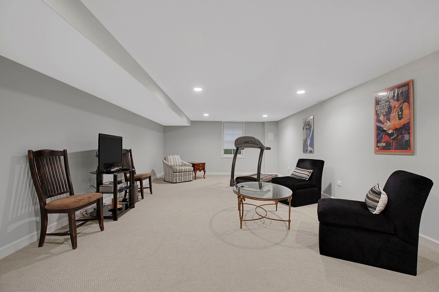This is a well-lit basement featuring neutral-toned carpeting and light gray walls. The space includes a seating area with dark armchairs, a glass-topped coffee table, and a television on a stand. A treadmill is also present, suggesting a multi-purpose use for the area, possibly as a recreational or exercise space.