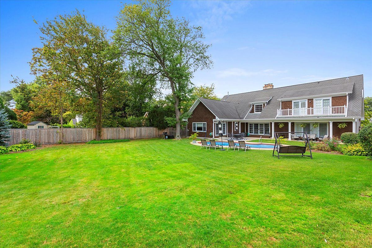 This image showcases a spacious backyard with a well-maintained lawn, mature trees, and a wooden fence. The rear of a two-story house is visible, featuring a balcony, patio, and a swimming pool with lounge chairs. A swing set adds a touch of leisure to the outdoor space, creating an inviting and private atmosphere.