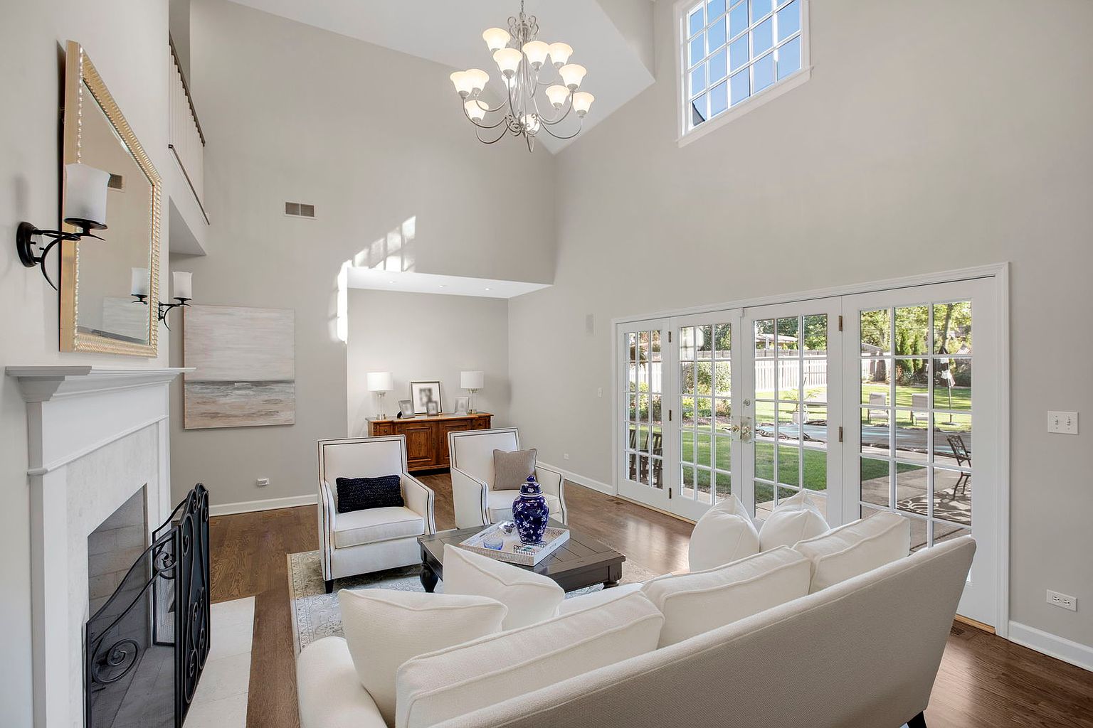This is an interior shot of a living room featuring a fireplace, two armchairs, and a white sofa. The room has high ceilings and large windows that let in natural light. The style is modern and elegant, with a focus on neutral colors and clean lines.