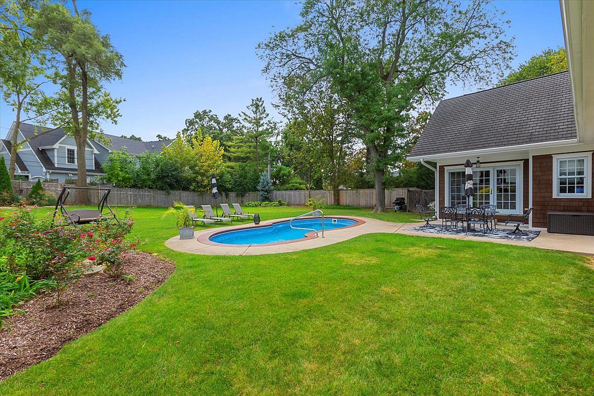 This image showcases a well-maintained backyard featuring a kidney-shaped pool, lush green lawn, and a patio area with outdoor seating. The yard is surrounded by mature trees and landscaping, providing privacy and a serene atmosphere. The house has brown siding and a dark roof, complementing the natural surroundings.