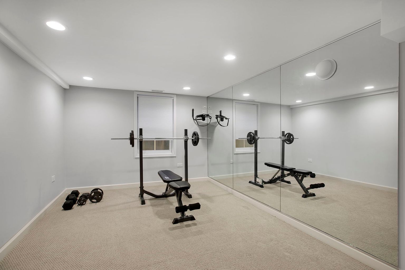 This is an interior shot of a home gym. The room features a weight bench with barbell, dumbbells, and a large mirror covering one wall, creating the illusion of a larger space. The walls are painted a neutral gray, and the floor is carpeted, providing a comfortable workout environment.