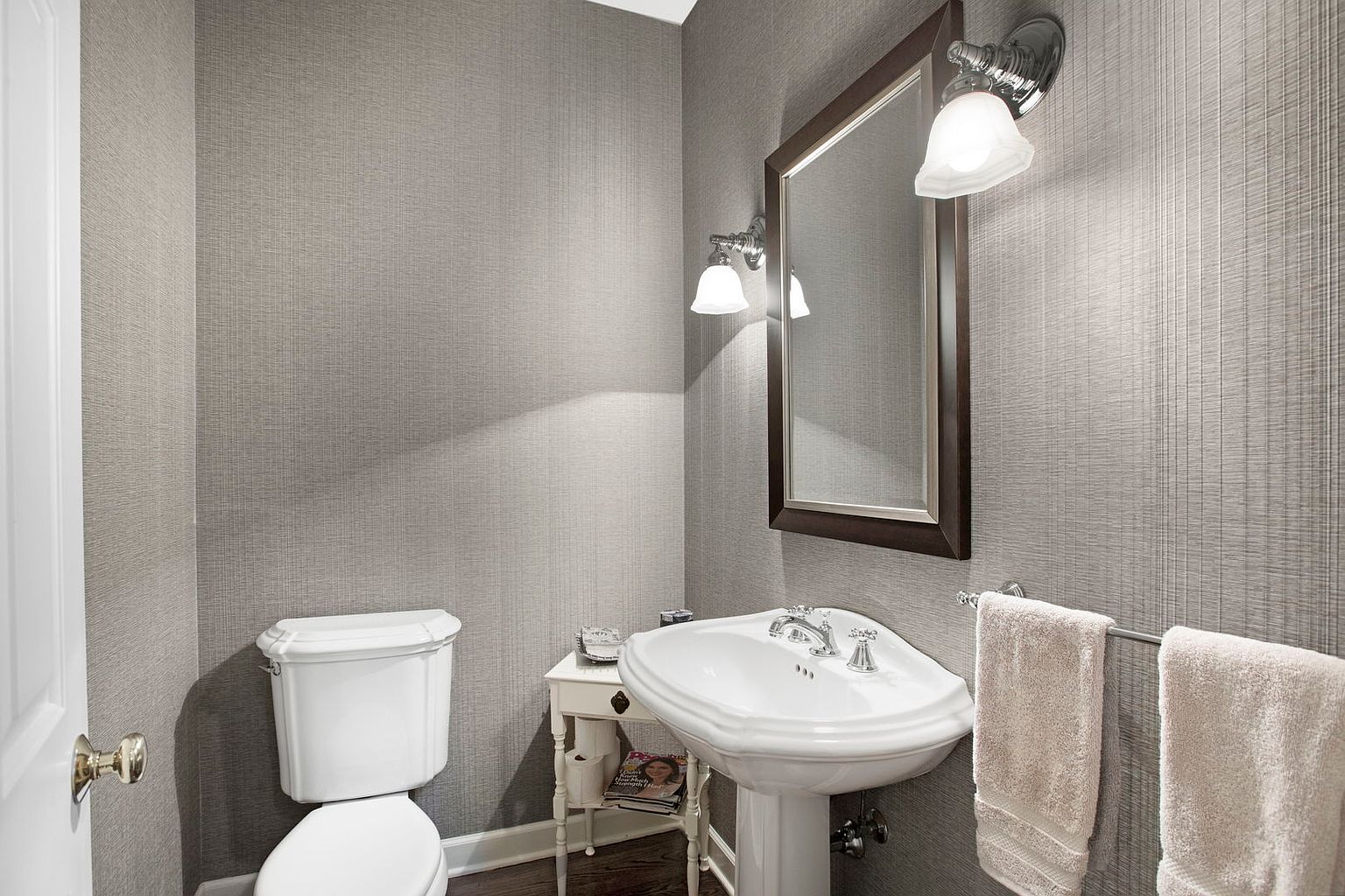 This is a well-lit, traditionally styled powder room. It features a pedestal sink, a framed mirror with sconce lighting, and a standard toilet. The walls are covered in a textured gray wallpaper, and the floor appears to be dark hardwood, creating a clean and classic look.