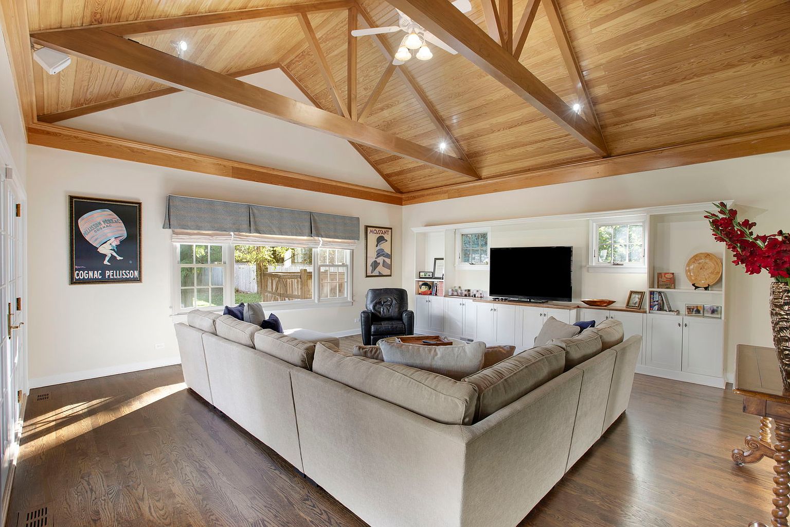 This is an interior shot of a living room featuring a high, vaulted ceiling with exposed wooden beams. A large sectional sofa anchors the space, facing a built-in entertainment center with a television. The room is well-lit with natural light from the windows, creating a warm and inviting atmosphere.