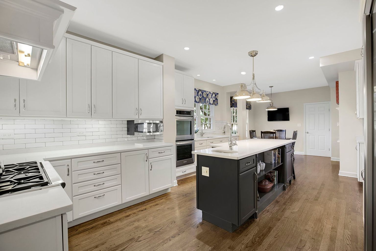 This is a bright and spacious kitchen featuring white cabinetry, subway tile backsplash, and stainless steel appliances. A dark-colored island with a white countertop provides ample workspace and storage. The hardwood floors add warmth to the space, and pendant lights illuminate the island, creating an inviting atmosphere.