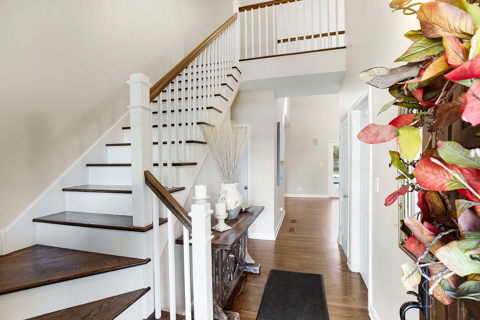 This interior shot showcases a well-lit hallway and staircase. The staircase features white risers and dark wood treads, complemented by a white banister with a wood handrail. A console table with decorative items sits adjacent to the stairs, and a dark rug lies on the hardwood floor, leading towards a doorway at the end of the hallway.
