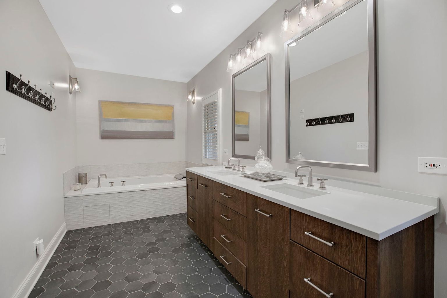 This is a well-lit primary bathroom featuring a double vanity with dark wood cabinets and a white countertop. Two large mirrors hang above the sinks, reflecting the room's neutral color palette. The floor is covered in a dark gray hexagonal tile, and a modern bathtub with textured white tiling is visible in the background, creating a luxurious and contemporary feel.